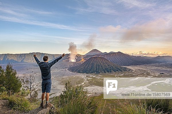 Junger Mann streckt Arme in die Luft  Vulkanlandschaft bei Sonnenuntergang  Blick in Tengger Caldera  rauchender Vulkan Gunung Bromo  vorne Mt. Batok  hinten Mt. Kursi  Mt. Gunung Semeru  Nationalpark Bromo-Tengger-Semeru  Java  Indonesien  Asien
