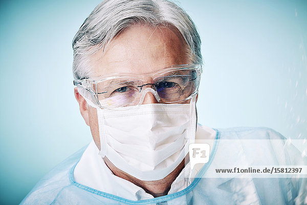 Grauhaariger Arzt mit Schutzhandschuhen und Schutzbrille blickt in Kamera