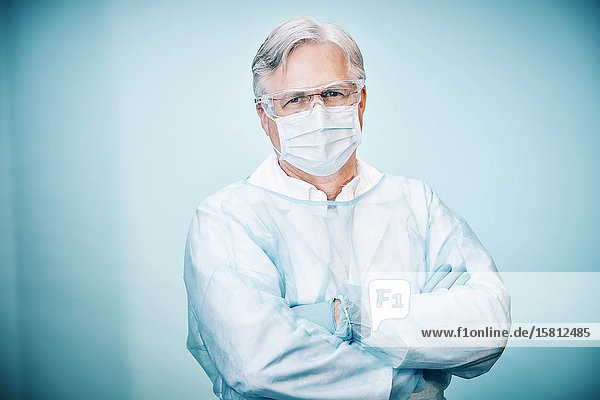 Arzt mit Schutzbrille und Mundschutz blickt mit verschränkten Armen in Kamera