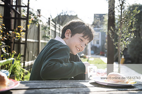 Lächelnder Junge in grünem Pullover  der an einem Tisch in einem Garten mit einem Imbiss sitzt.