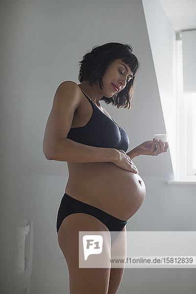Schwangere Frau in BH und Höschen trägt Feuchtigkeitscreme auf den Bauch auf