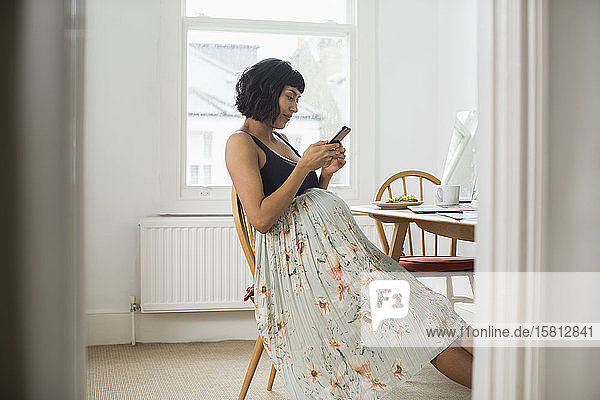 Schwangere Frau im geblümten Kleid benutzt ihr Smartphone am Tisch
