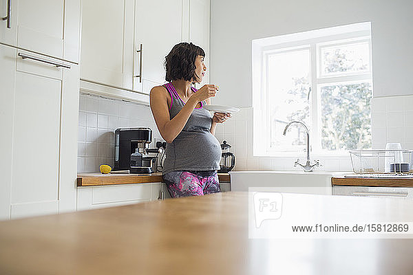 Nachdenkliche schwangere Frau  die in der Küche isst und aus dem Fenster schaut