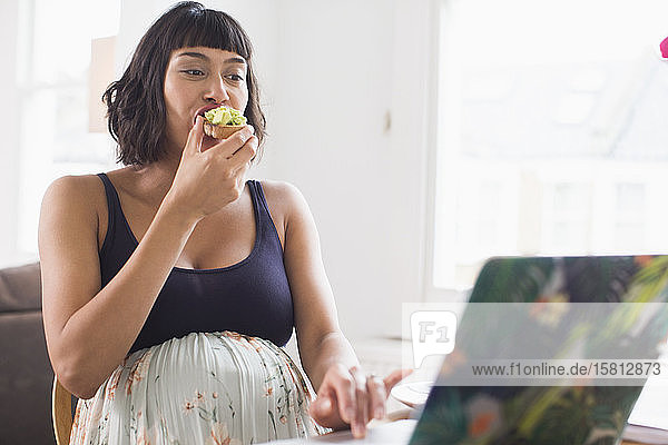 Pregnant woman eating avocado toast at laptop