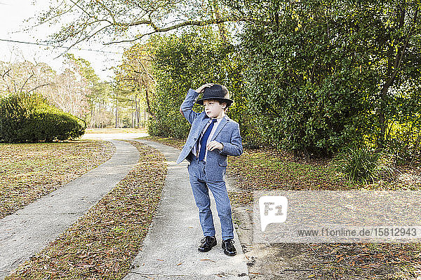 A six year old boy dressed in suit and wearing fedora  in driveway