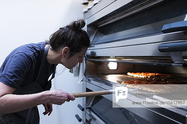 Frau mit Schürze steht in einer handwerklichen Bäckerei und schiebt Pizza in den Ofen.
