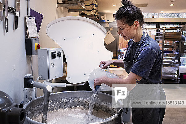 Frau mit Schürze steht in einer handwerklichen Bäckerei und gießt Wasser in einen industriellen Mixer.