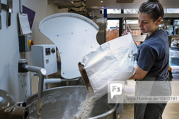 Frau mit Schürze steht in einer handwerklichen Bäckerei und schüttet Mehl in einen industriellen Mixer.