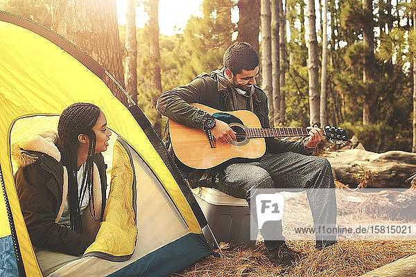 Young couple playing guitar at tent at campsite in woods