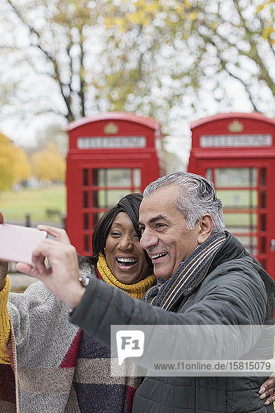 Glückliches älteres Paar  das ein Selfie vor einer roten Telefonzelle im Herbstpark macht