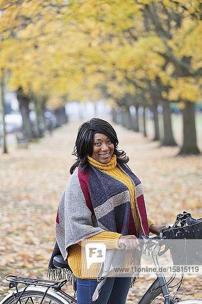 Porträt lächelnde Frau mit Fahrrad unter Blättern und Bäumen im Herbst Park