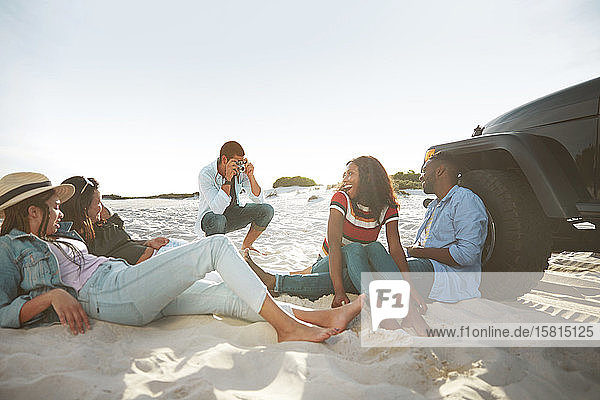 Junger Mann mit Digitalkamera  der seine Freunde am sonnigen Strand fotografiert