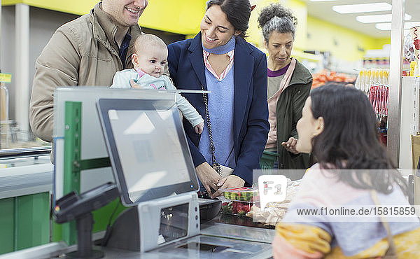 Freundliche Kassiererin im Gespräch mit und Hilfe für ein Paar mit Baby an der Supermarktkasse