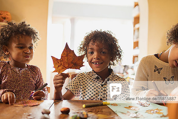 Portrait glücklicher Junge mit Herbstblatt beim Basteln mit Schwestern am Tisch