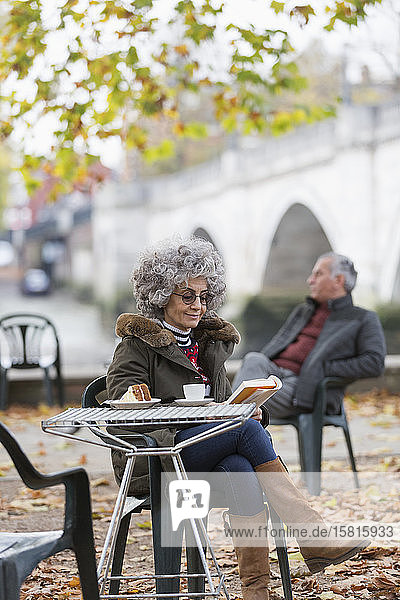 Aktive Seniorin liest ein Buch und genießt Kaffee und Kuchen im Herbstparkcafé