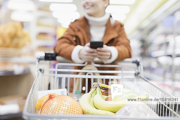 Frau mit Smartphone schiebt Einkaufswagen im Supermarkt