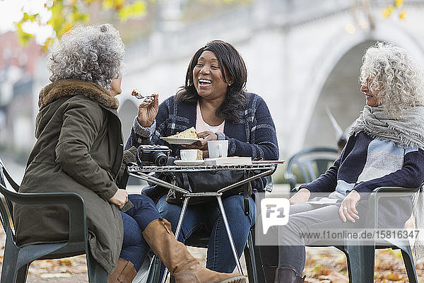 Laughing active senior women friends sharing dessert at autumn park cafe