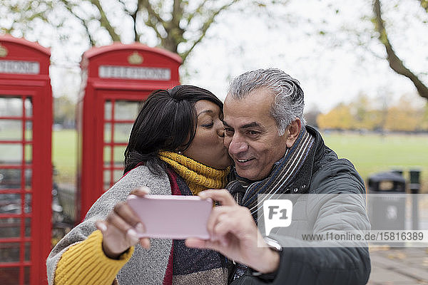 Älteres Paar  das sich küsst und ein Selfie im Park vor einer roten Telefonzelle macht