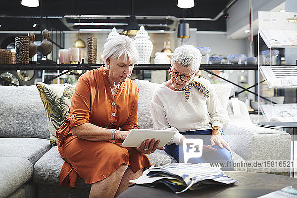 Ältere Frau mit digitalem Tablet betrachtet Stoffmuster auf einem Sofa in einem Möbelhaus