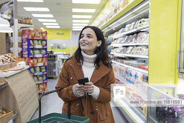 Lächelnde junge Frau mit Smartphone beim Lebensmitteleinkauf im Supermarkt