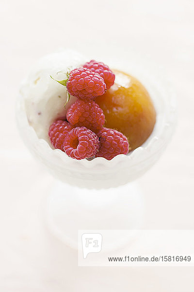 Dessertschale aus Milchglas mit einer Kugel Vanilleeis  einem pochierten ganzen Pfirsich und frisch gepflückten Himbeeren