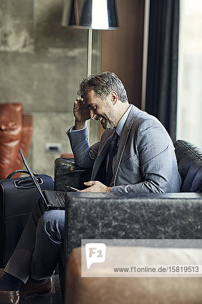 Smiling businessman using laptop and smartphone in hotel lobby