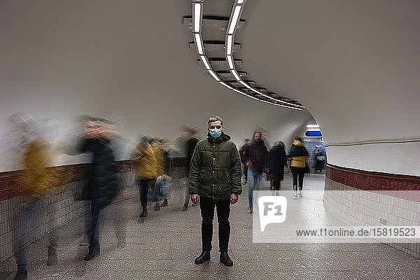 Young man with face mask standing isolated in subway underpass  with people moving around him