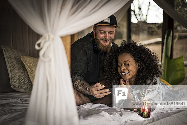 Glückliches Paar im Bett in einer Lodge mit Smartphone und frischem Eistee