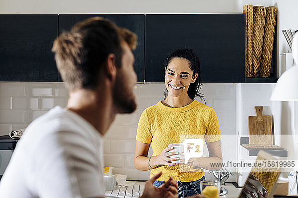 Portrait of young woman smiling at boyfriend with laptop in the kitchen