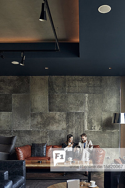 Businessman and businesswoman sitting on couch in hotel lobby sharing smartphone
