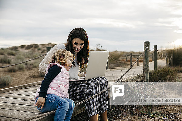 Mother with laptop and daughter on a boardwalk in the countryside