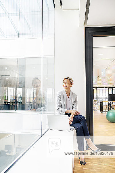 Blond businesswoman with laptop  sitting on windowsill in office building