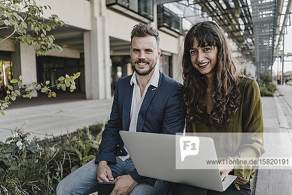 Porträt eines lächelnden Geschäftsmannes und einer lässigen Geschäftsfrau  die einen Laptop im Freien benutzen