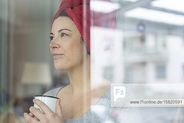 Bildnis einer Frau mit in ein Handtuch gewickeltem Kopf die hinter einer Fensterscheibe einen Kaffee trinkt