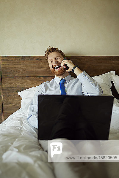 Geschäftsmann im Hotelzimmer auf dem Bett liegend und am Telefon lachend