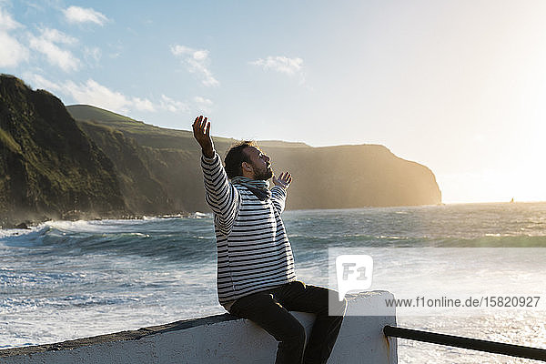 Man sitting at the coast at sunset with outstretched arms  Sao Miguel Island  Azores  Portugal
