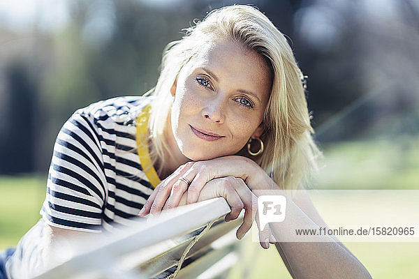 Blond lächelnde Frau  die auf einer Bank sitzt und in die Kamera schaut