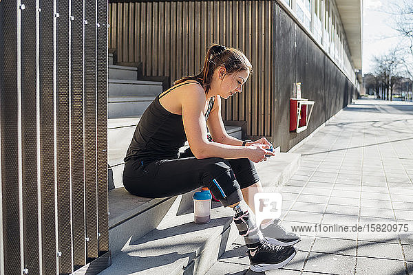 Sportliche junge Frau mit Beinprothese sitzt auf einer Treppe in der Stadt und benutzt ein Smartphone