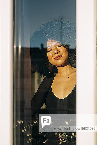 Porträt einer jungen Frau mit lockigem Haar hinter einer Fensterscheibe  die den Sonnenschein genießt