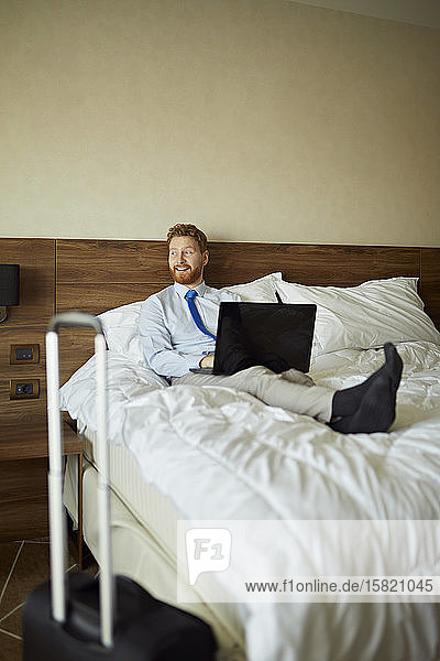 Businessman lying on bed in hotel room using laptop