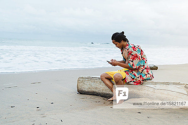 Junger Mann sitzt mit seinem Handy auf totem Holz am Strand  Insel Borneo  Malaysia