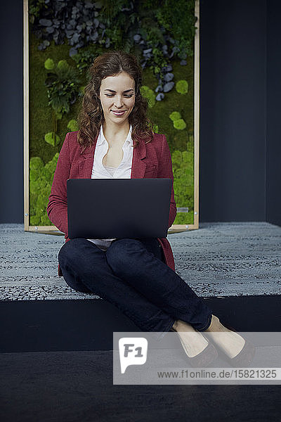 Lächelnde Geschäftsfrau sitzt in grünem Büro mit Laptop auf dem Boden