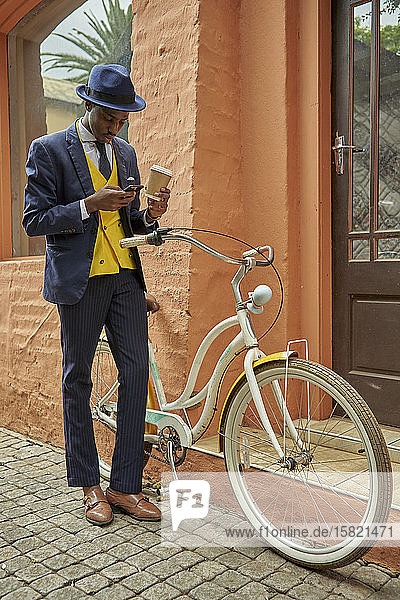 Modischer junger Geschäftsmann mit Fahrrad im altmodischen Anzug prüft sein Telefon