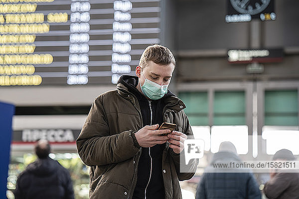 Junger Mann mit Gesichtsmaske am Bahnhof in der Stadt  der ein Smartphone benutzt