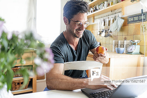Happy man sitting at table in kitchen using laptop