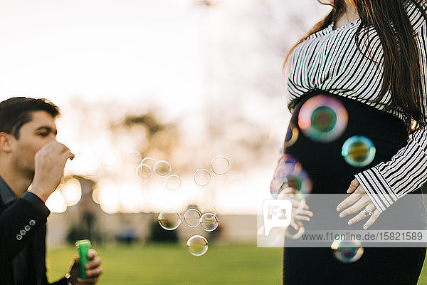 Expectant parents blowing soap bubbles in a park at sunset