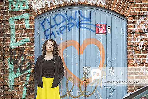 Bildnis einer brünetten Frau  die vor einer Ziegelmauer mit einem alten Tor und Graffiti steht
