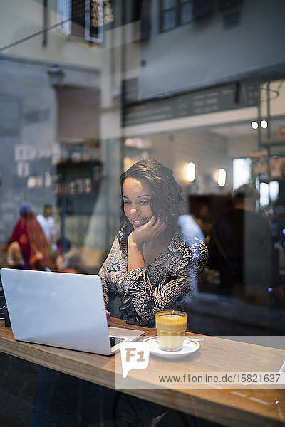 Junge Frau mit Laptop in einem Café hinter einer Fensterscheibe