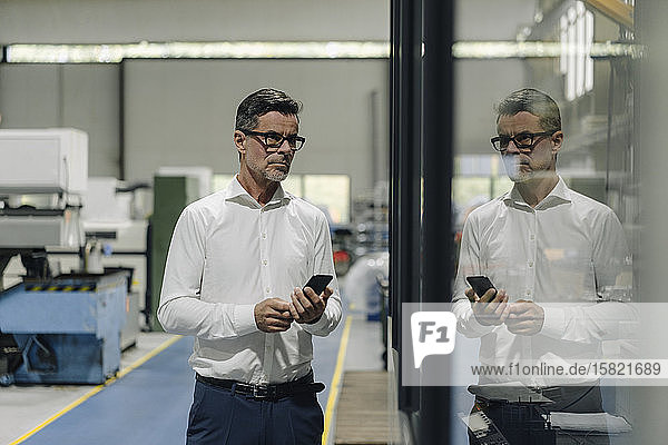Mature businessman with cell phone in a factory reflected in glass pane