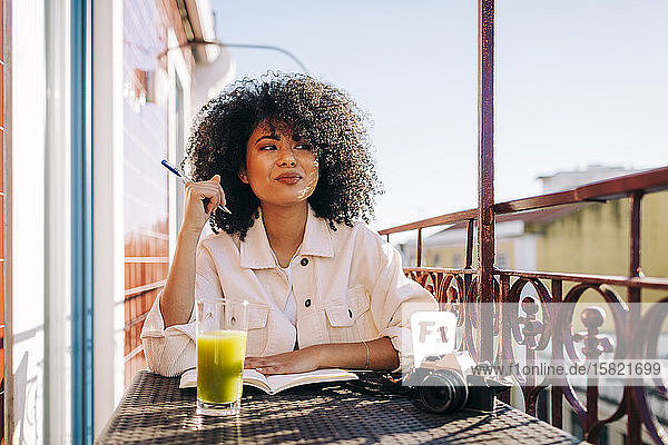 Lächelnde junge Frau mit lockigem Haar sitzt am Tisch auf dem Balkon mit Notebook  Smoothie und Kamera